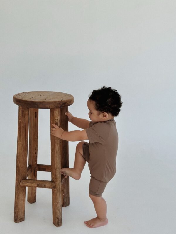 A baby is standing on a stool in front of a white background wearing the Essentials Range - Zimmi Onesie - Tort.