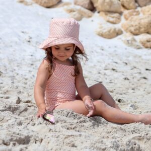 A little girl sitting in the Retro Wave By Ina - Mara One-Piece - Marigold Stripe with a hat on.