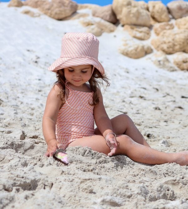 A little girl sitting in the Retro Wave By Ina - Mara One-Piece - Marigold Stripe with a hat on.