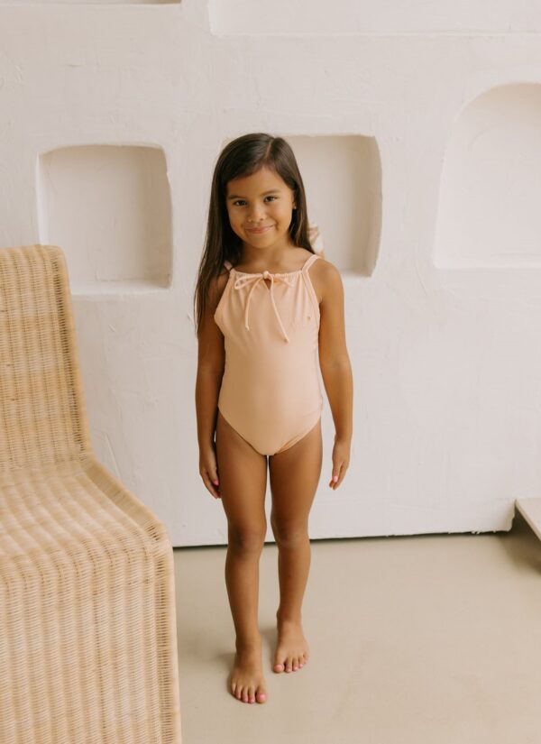 A young girl in an Aurelia One-Piece - Peach Blossom swimsuit standing in a light-colored room with a wicker chair to the side.