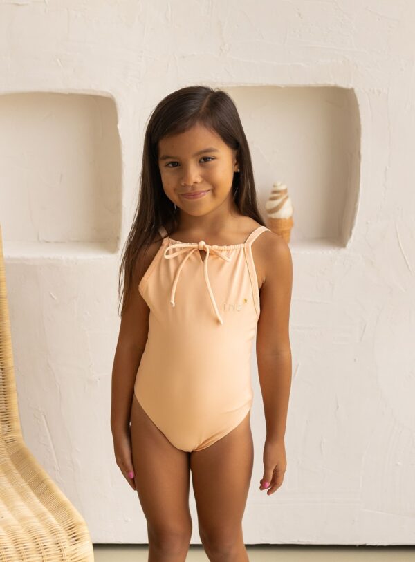 Young girl in an Aurelia One-Piece - Peach Blossom swimsuit standing beside a white wall, holding an ice cream.