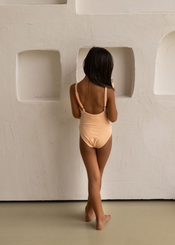 A woman in an Aurelia One-Piece - Peach Blossom swimsuit standing in front of a textured wall with geometric cut-outs.