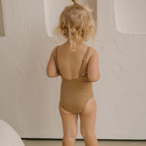 A toddler wearing an Aurelia One-Piece - Warm Pecan with a topknot hairstyle stands facing a textured wall.