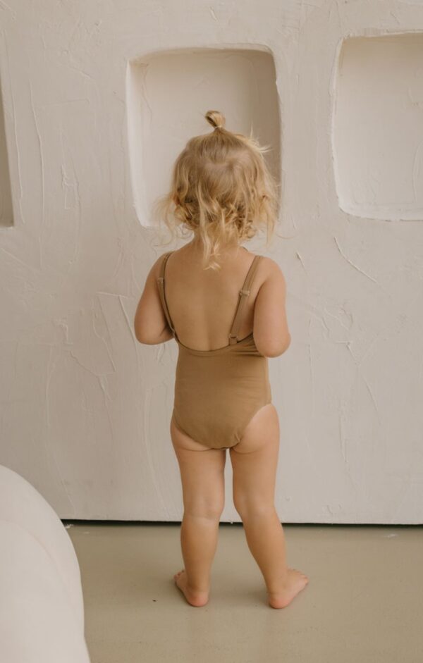 A toddler wearing an Aurelia One-Piece - Warm Pecan with a topknot hairstyle stands facing a textured wall.