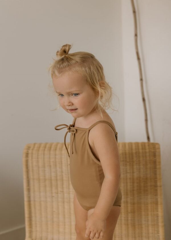 A young child with a topknot hairstyle standing beside a wicker chair, wearing the Aurelia One-Piece - Warm Pecan swimsuit and appearing slightly pensive.