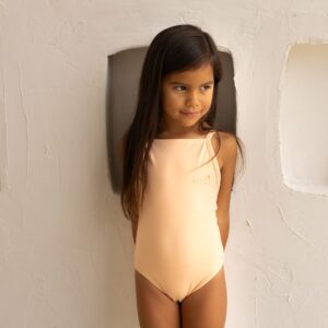 Young girl posing in a Aurelia One-Piece - Peach Blossom swimsuit against a textured wall with niche-like openings.
