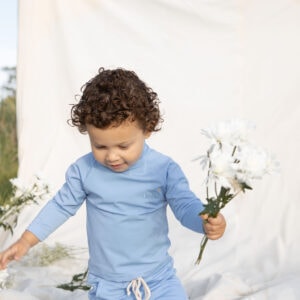 A young child with curly hair holding flowers while walking on a draped Nella Rash Shirt - Powder Sky outdoors.