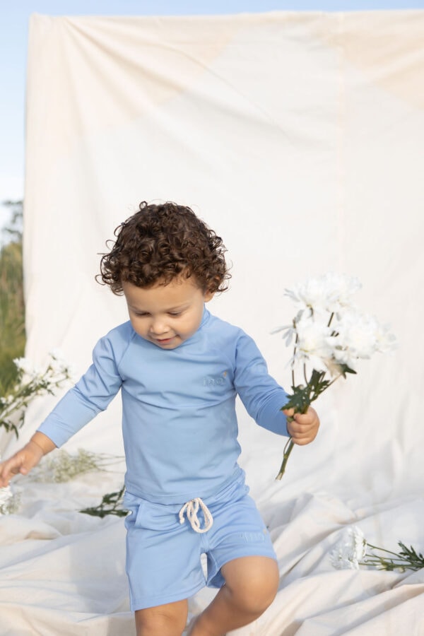A young child with curly hair holding flowers while walking on a draped Nella Rash Shirt - Powder Sky outdoors.