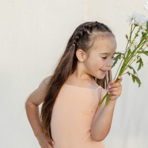 A young girl in a Mara One-Piece - Peach Blossom leotard smelling white flowers against a light background.