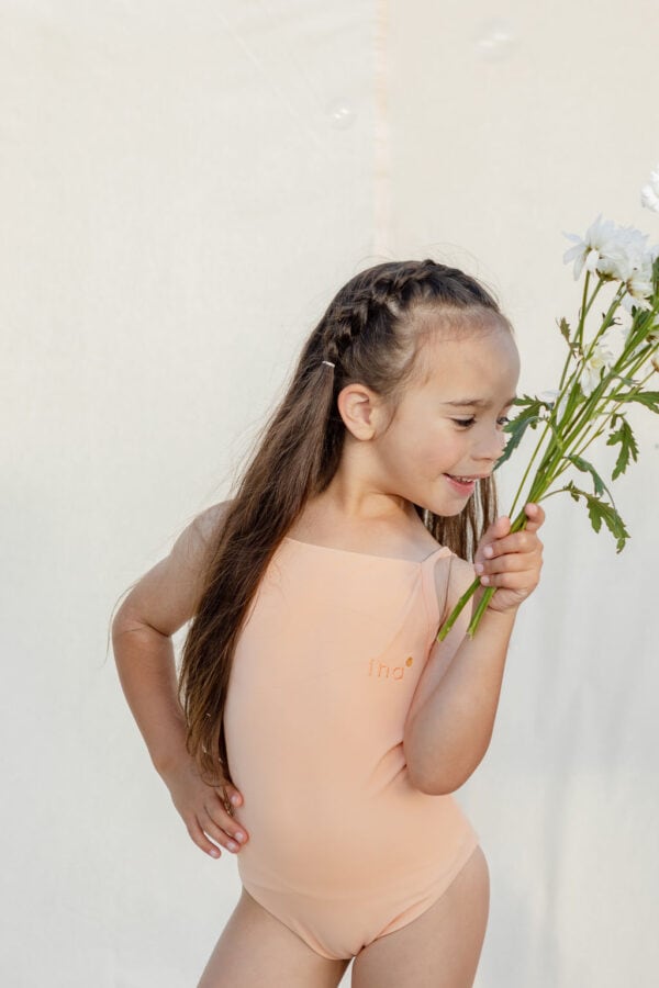 A young girl in a Mara One-Piece - Peach Blossom leotard smelling white flowers against a light background.