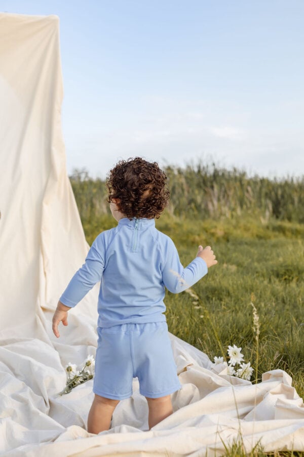 Toddler in Nella Rash Shirt - Powder Sky standing next to white cloth backdrop in a field.