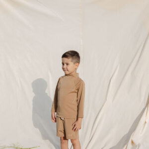 Young boy in Amias Trunks - Warm Pecan attire standing against a cream backdrop outdoors.