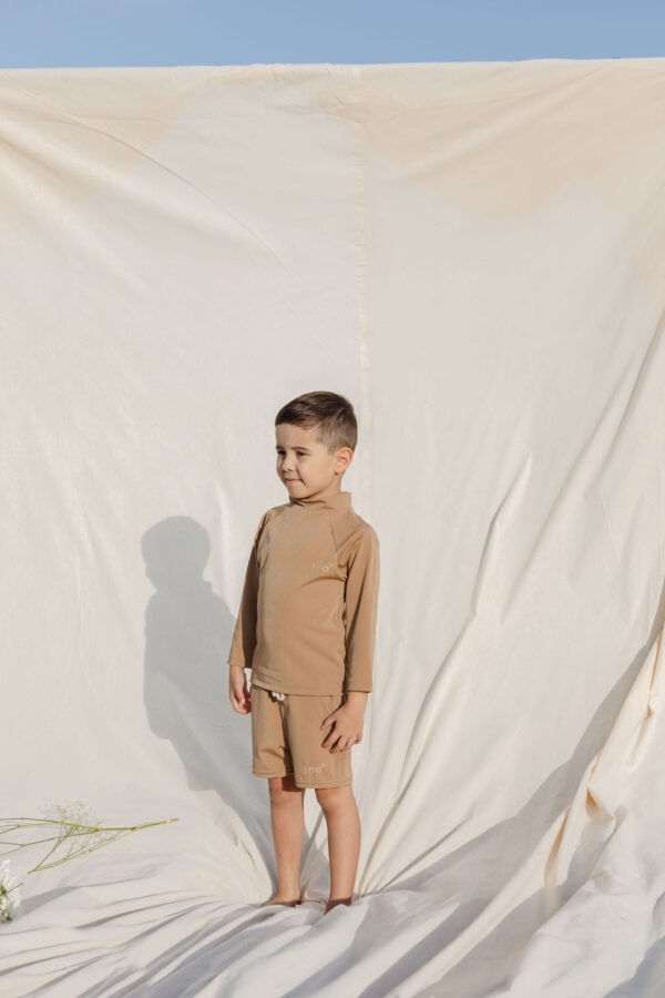 Young boy in Amias Trunks - Warm Pecan attire standing against a cream backdrop outdoors.