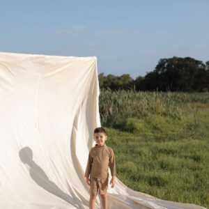 A child stands next to a draped white fabric in an open field wearing Amias Trunks - Warm Pecan.