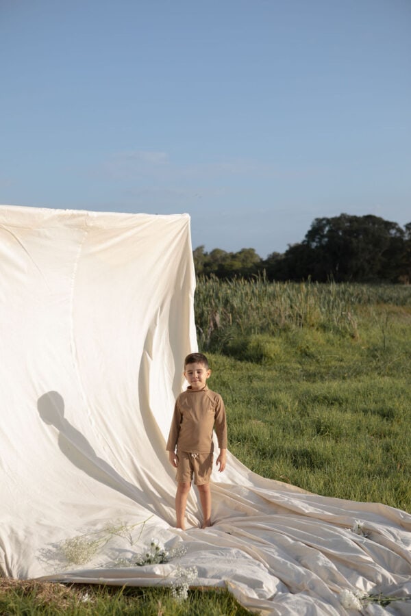 A child stands next to a draped white fabric in an open field wearing Amias Trunks - Warm Pecan.