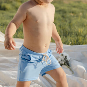 A toddler in Amias Trunks - Powder Sky standing on a white blanket in a field.