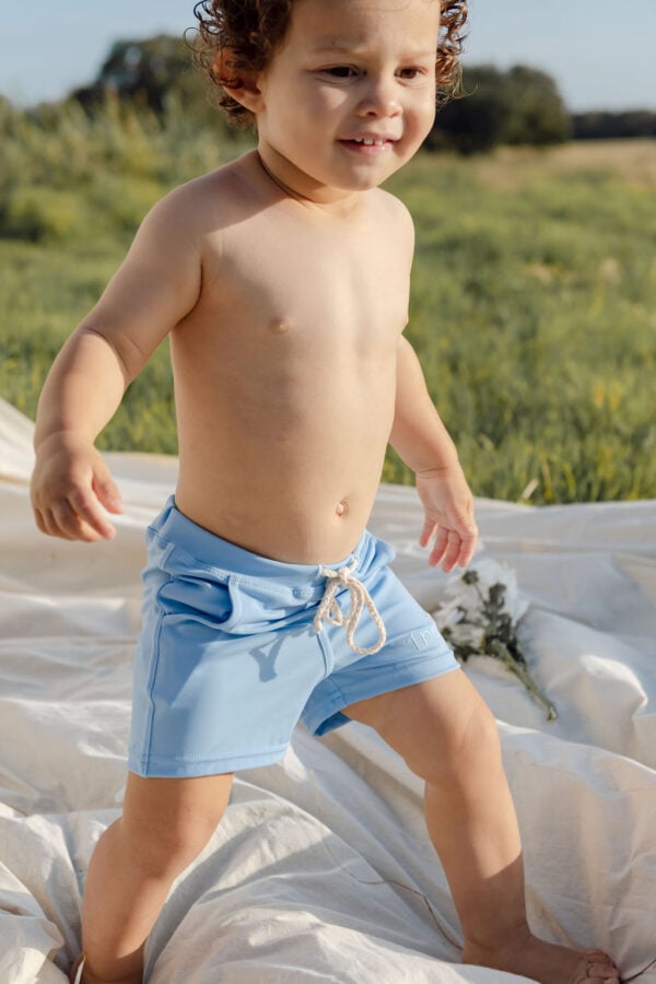 A toddler in Amias Trunks - Powder Sky standing on a white blanket in a field.