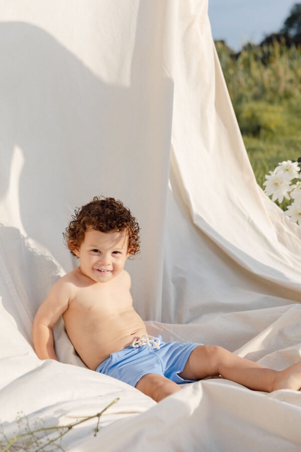 Toddler with curly hair smiling while sitting on a white cloth outdoors wearing Mesa Trunks - Powder Sky.