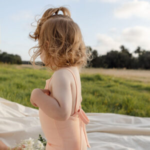 A young child in a Mara One-Piece - Peach Blossom swimsuit standing on a blanket outdoors with flowers nearby.