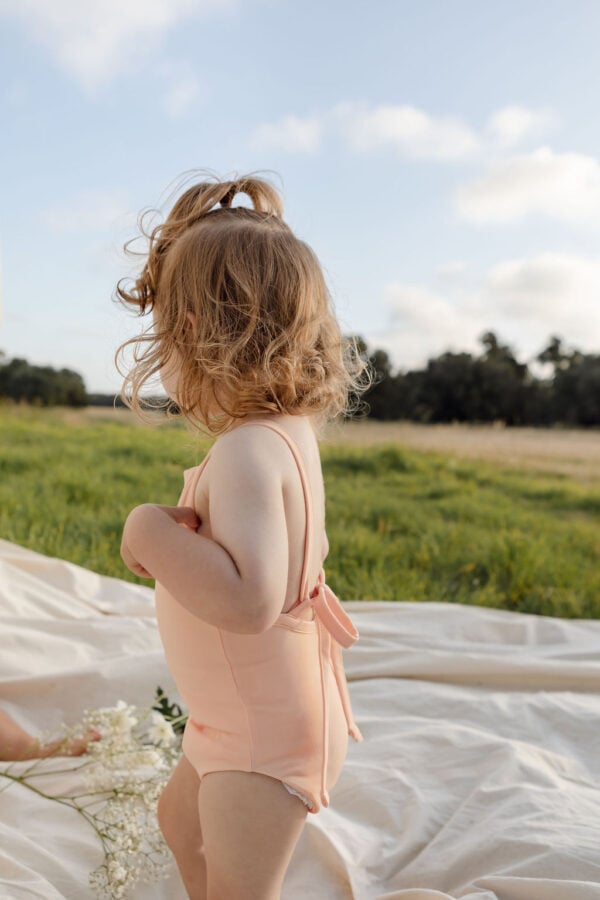 A young child in a Mara One-Piece - Peach Blossom swimsuit standing on a blanket outdoors with flowers nearby.