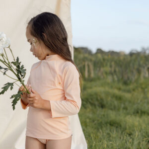 A young girl in a Nella Rash Shirt - Peach Blossom and long-sleeve top holding a flower while standing in a field beside a draped white cloth.