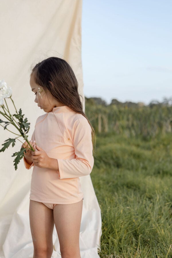 A young girl in a Nella Rash Shirt - Peach Blossom and long-sleeve top holding a flower while standing in a field beside a draped white cloth.