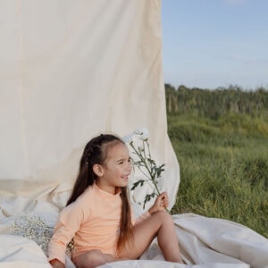 A young girl sitting on a blanket outdoors, holding a Nella Rash Shirt - Peach Blossom, with a draped white fabric in the background.
