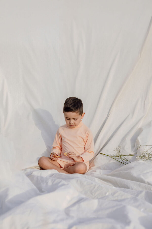 A child sitting cross-legged on a white Nella Rash Shirt - Peach Blossom, looking down with a pensive expression.