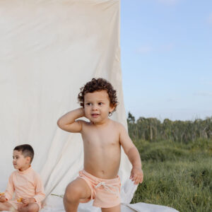 A young child standing with one hand on their head against a white backdrop in a natural setting, with another child seated in the background wearing Mesa Trunks - Peach Blossom.