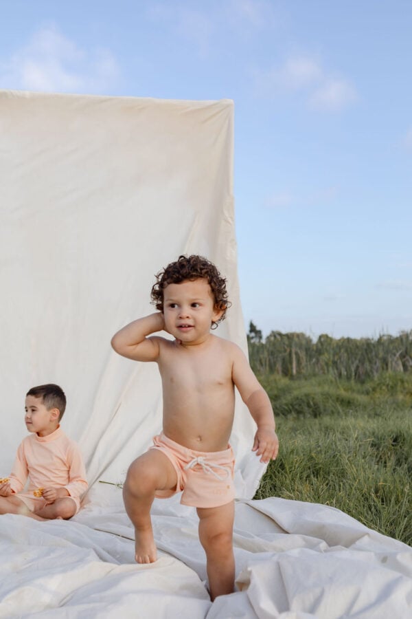 A young child standing with one hand on their head against a white backdrop in a natural setting, with another child seated in the background wearing Mesa Trunks - Peach Blossom.