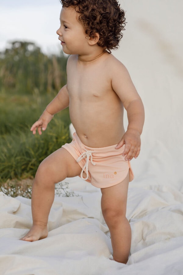 A toddler with curly hair stands on a blanket in a grassy area, wearing Mesa Trunks in Peach Blossom.