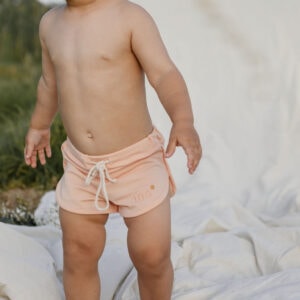 A toddler in Mesa Trunks - Peach Blossom standing on a white blanket outdoors.