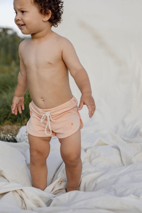 A toddler in Mesa Trunks - Peach Blossom standing on a white blanket outdoors.
