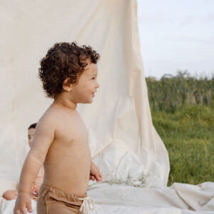 A joyful toddler with curly hair walking on a blanket outdoors with a Mesa Trunks - Warm Pecan backdrop.
