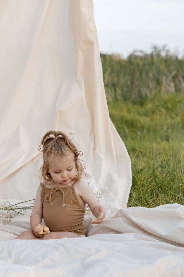 A toddler sitting on a white cloth outdoors, holding an Aurelia One-Piece - Warm Pecan, with a cream backdrop fluttering in the background.