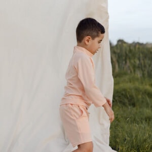 Young boy in Amias Trunks - Peach Blossom outfit stepping onto a white cloth outdoors.