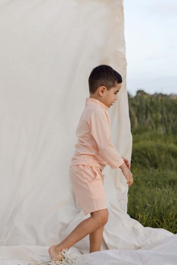 Young boy in Amias Trunks - Peach Blossom outfit stepping onto a white cloth outdoors.