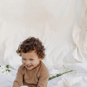 A smiling child with curly hair sits on a Nella Rash Shirt - Warm Pecan fabric background, near white flowers.
