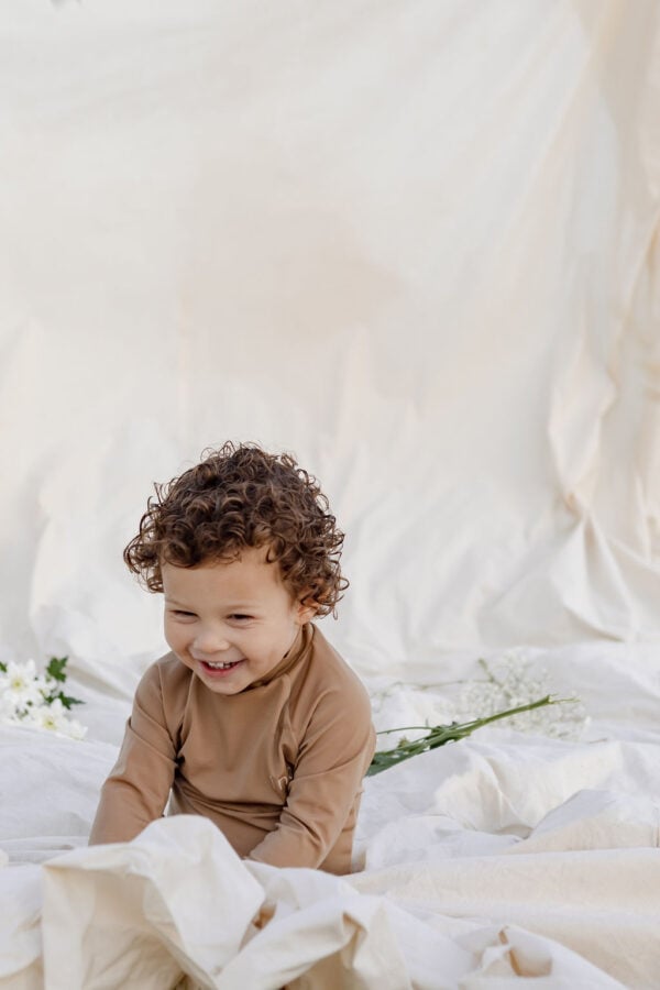 A smiling child with curly hair sits on a Nella Rash Shirt - Warm Pecan fabric background, near white flowers.