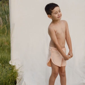 A young boy standing in front of a plain backdrop, squinting and wearing Amias Trunks - Peach Blossom.