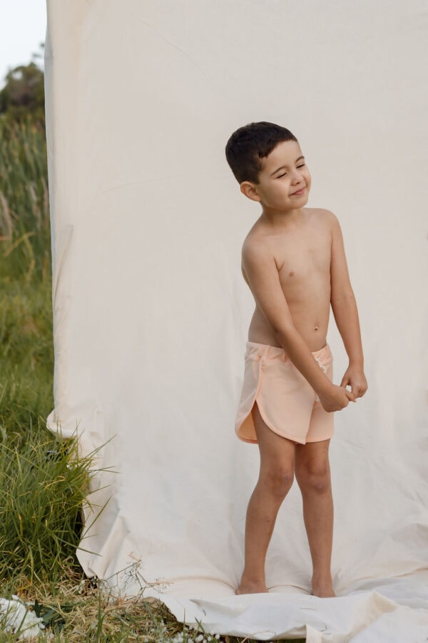 A young boy standing in front of a plain backdrop, squinting and wearing Amias Trunks - Peach Blossom.