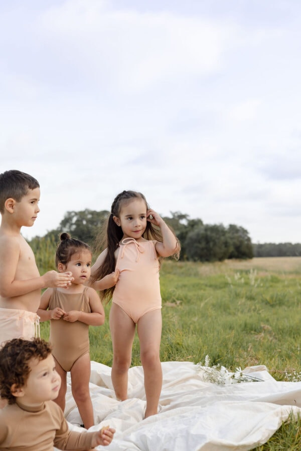 Four young children in coordinating Aurelia One-Piece - Peach Blossom outfits standing outdoors on a blanket with a natural landscape in the background.