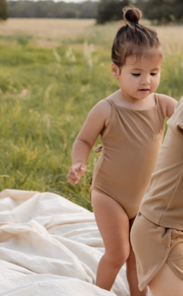 Toddler in a Mara One-Piece - Warm Pecan outfit with an adult on a blanket in a grassy field.