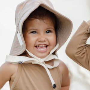 A cheerful young child wearing a beige sun hat and swimsuit with a hood smiling brightly in the Mara One-Piece - Warm Pecan.