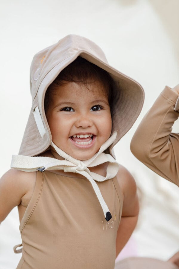 A cheerful young child wearing a beige sun hat and swimsuit with a hood smiling brightly in the Mara One-Piece - Warm Pecan.