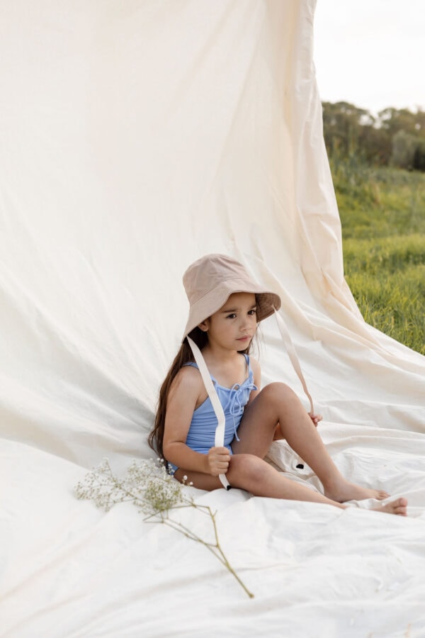 A young child in an Aurelia One-Piece - Powder Sky swimsuit and sunhat sitting under a draped cloth outdoors.