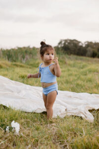 A toddler in a Luna Bikini - Powder Sky standing on grass with a curious expression, holding a flower petal.