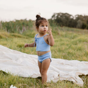 A toddler in a Luna Bikini - Powder Sky standing on grass with a curious expression, holding a flower petal.