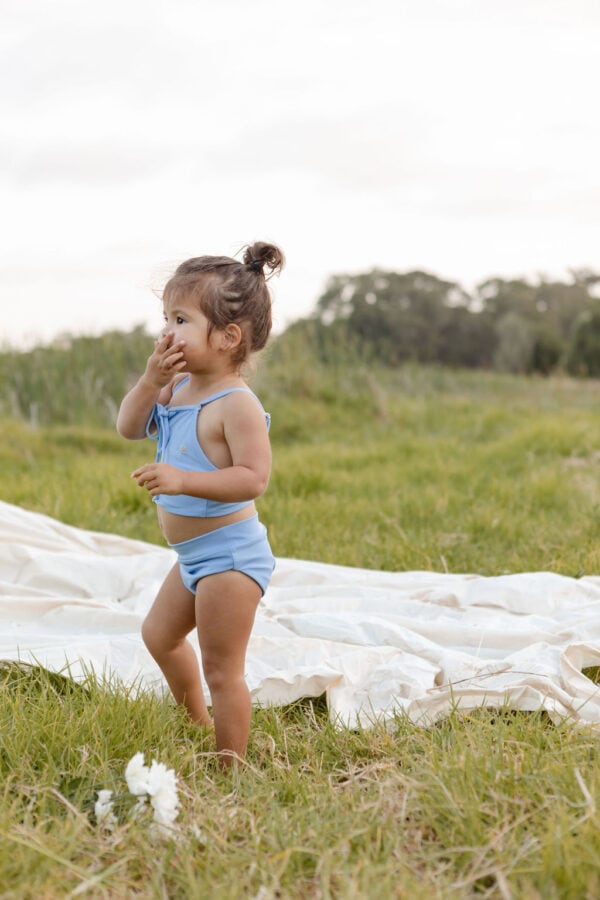 A toddler in a Luna Bikini - Powder Sky outfit standing in a field with a curious expression, touching their mouth with a hand.