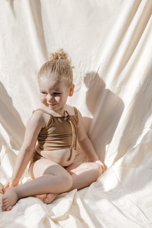 Toddler in a Luna Bikini - Warm Pecan sitting on a cream fabric with sunlight casting shadows.